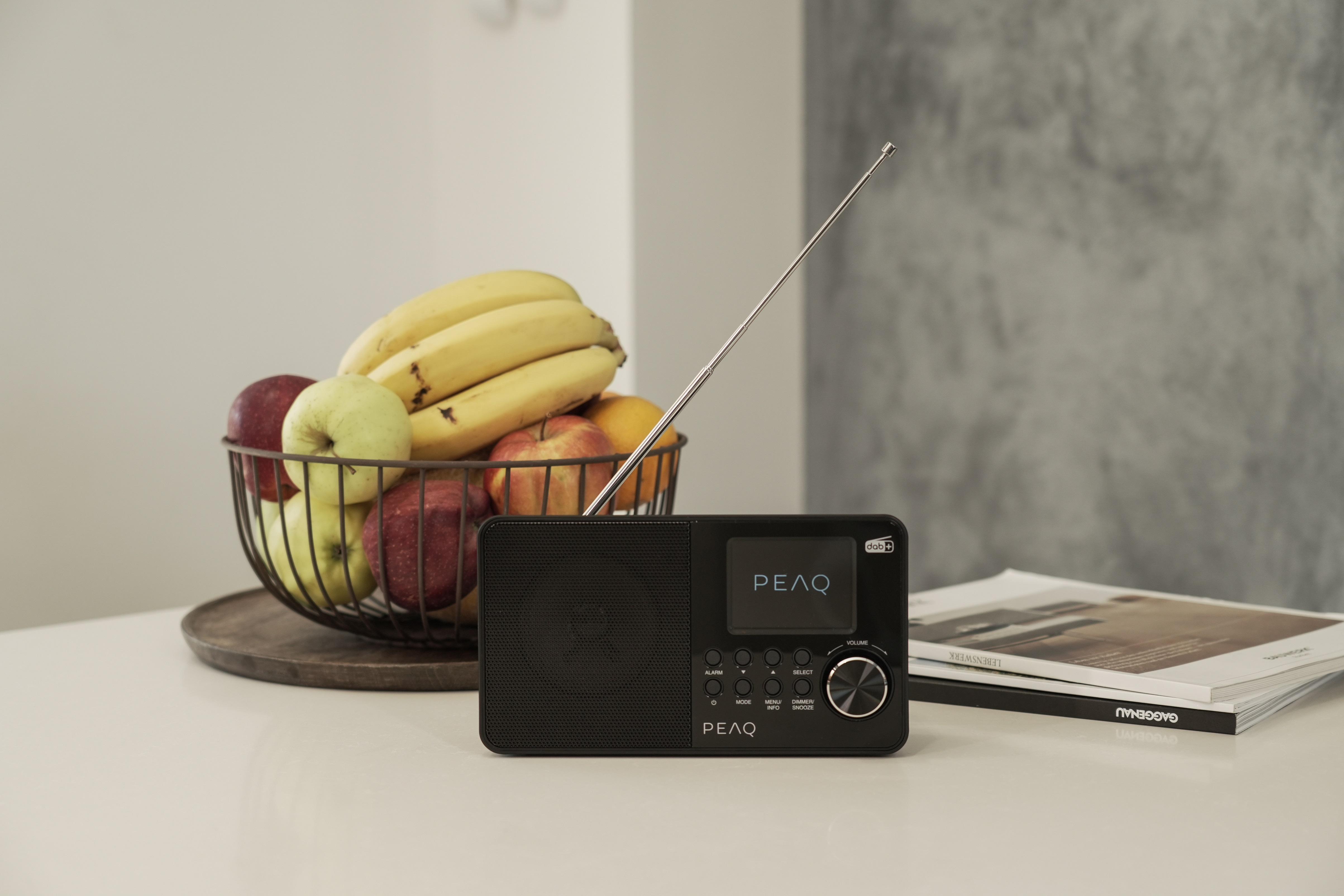 Czarny radio z metalową anteną, koszem owoców i czasopismami na stole.