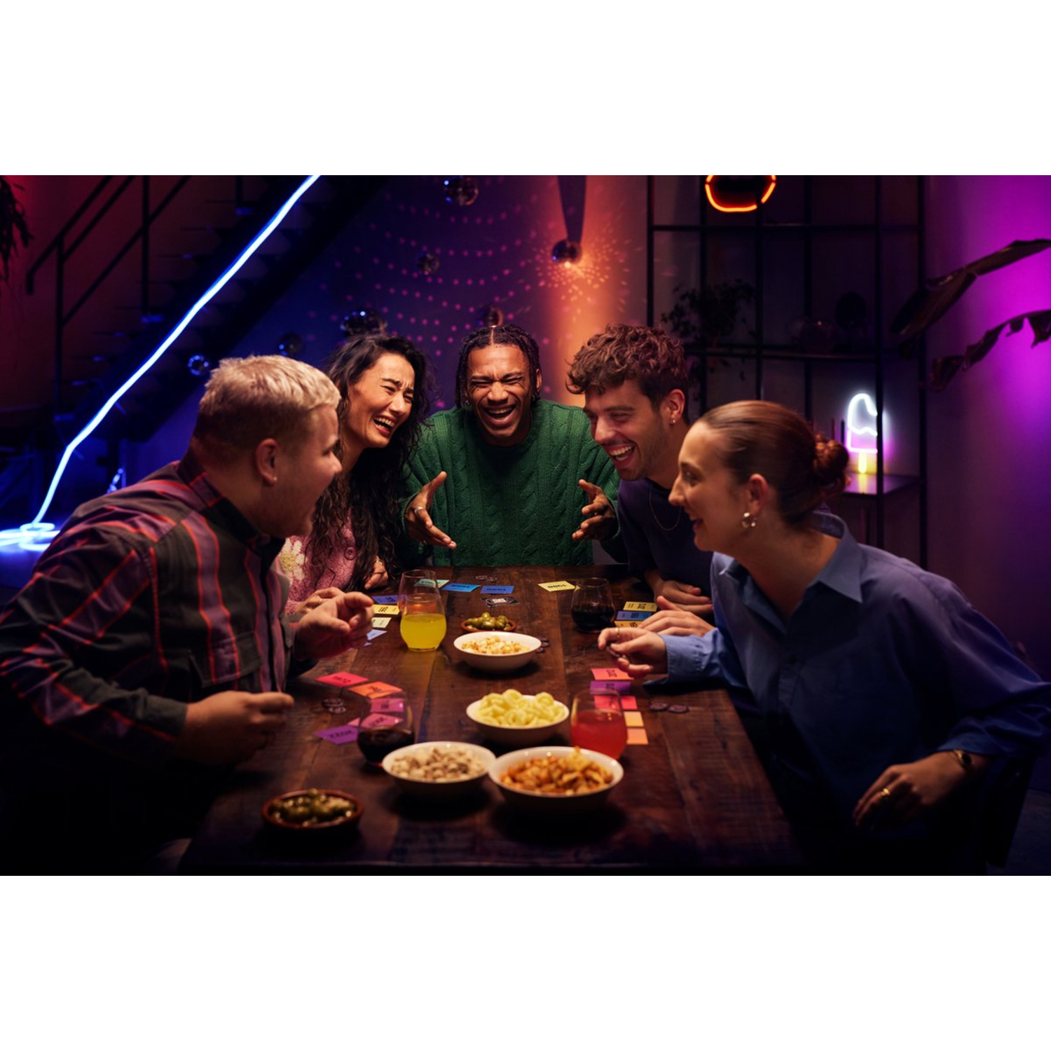 Vijf mensen verzameld rond een houten tafel, lachen en spelen een bordspel met snacks in de buurt.