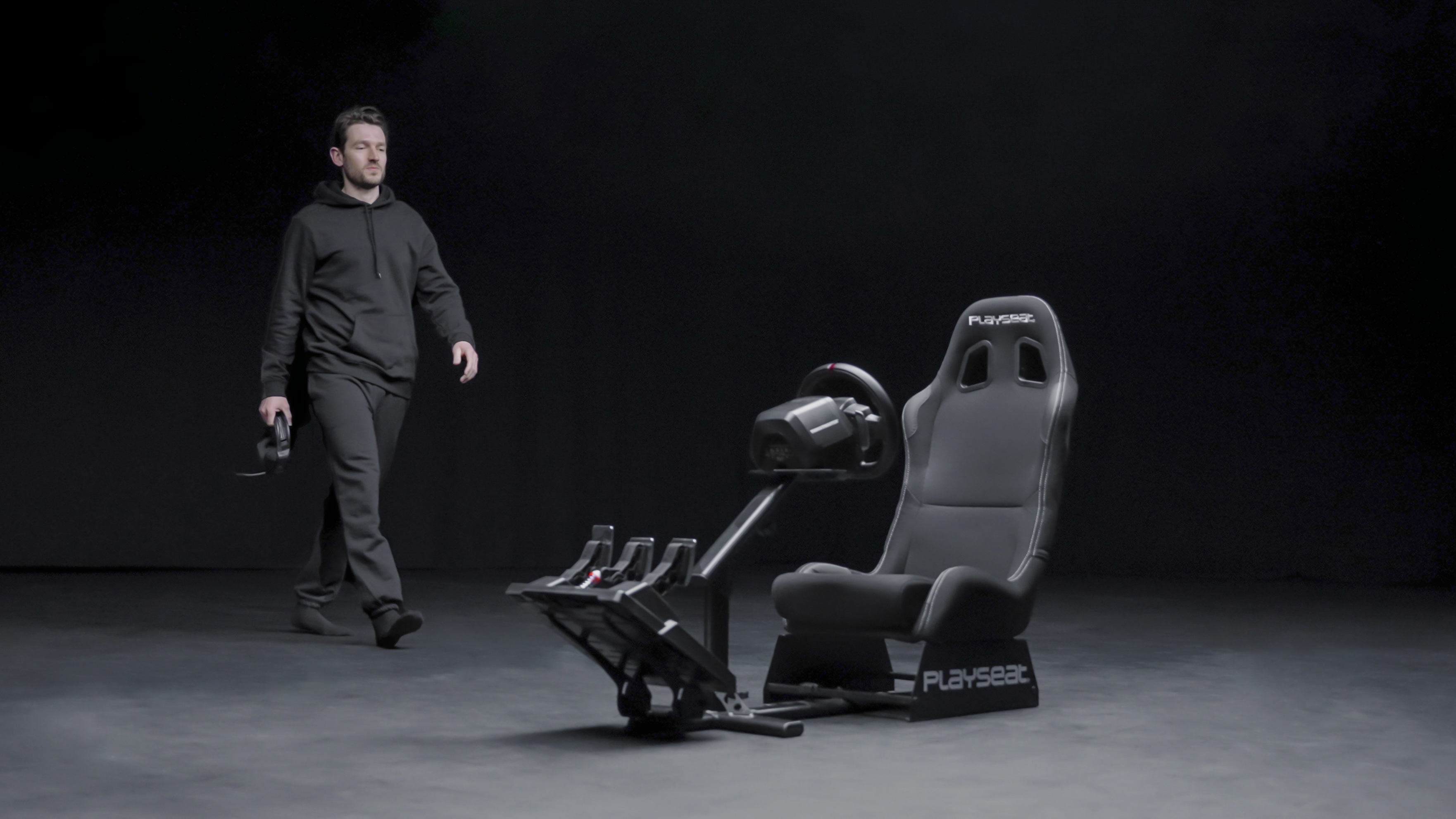 Mann geht auf einen Rennsitz mit Lenkrad und Pedalen zu. Dunkler Hintergrund.