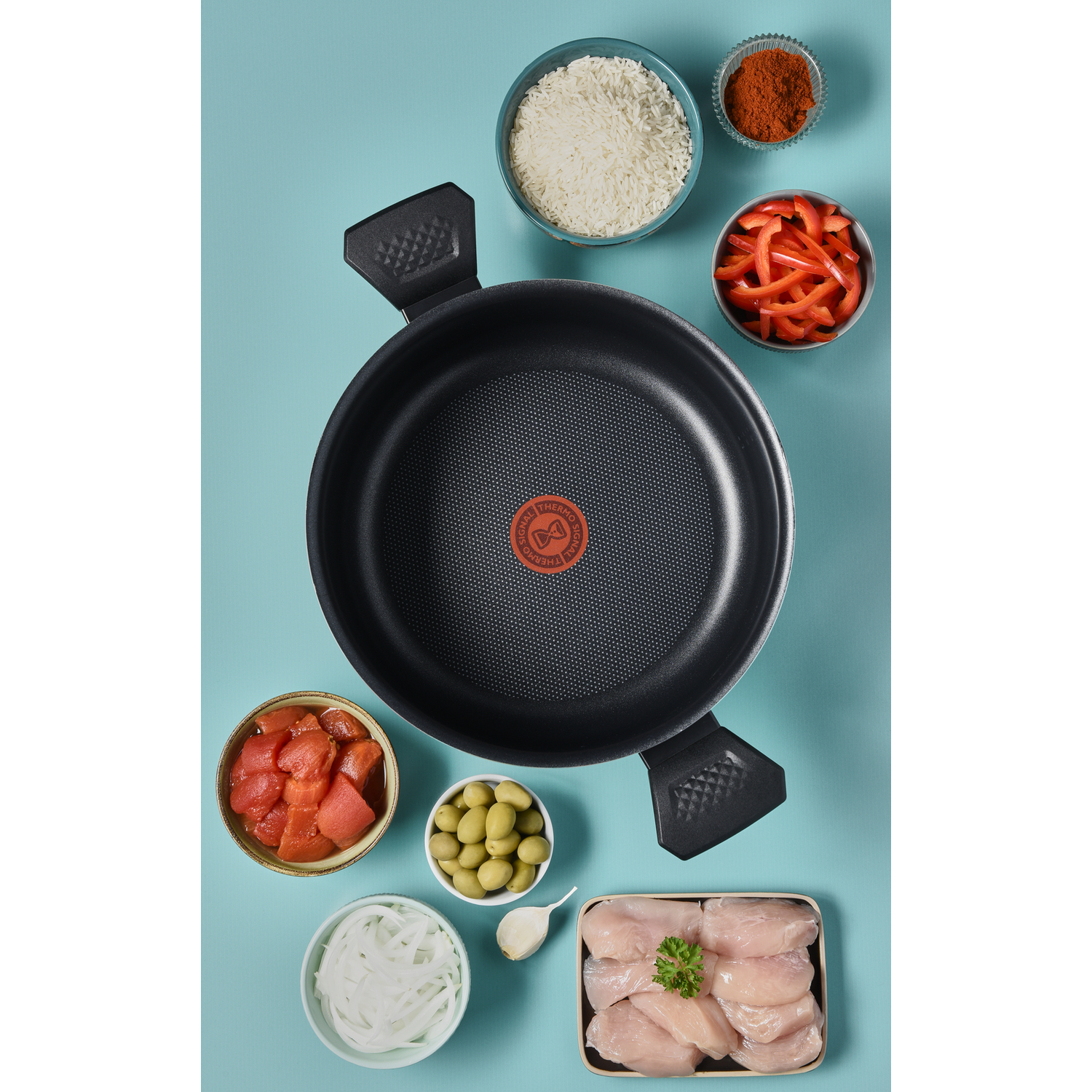 A black frying pan surrounded by ingredients like tomatoes, olives, chicken, and rice on a blue surface.
