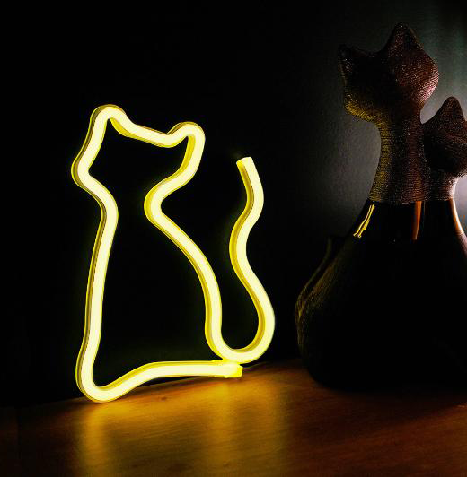 Neon macska körvonala sárga fénnyel, egy fekete macska alakú dekoráció mellett, sötét háttéren.