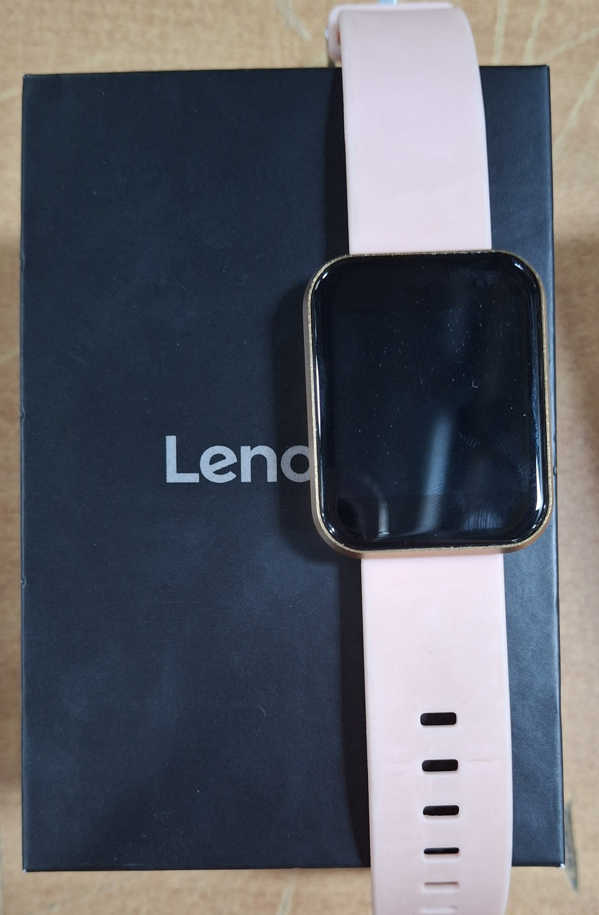 Różowy smartwatch ze złotą ramką leży na czarnym pudełku.