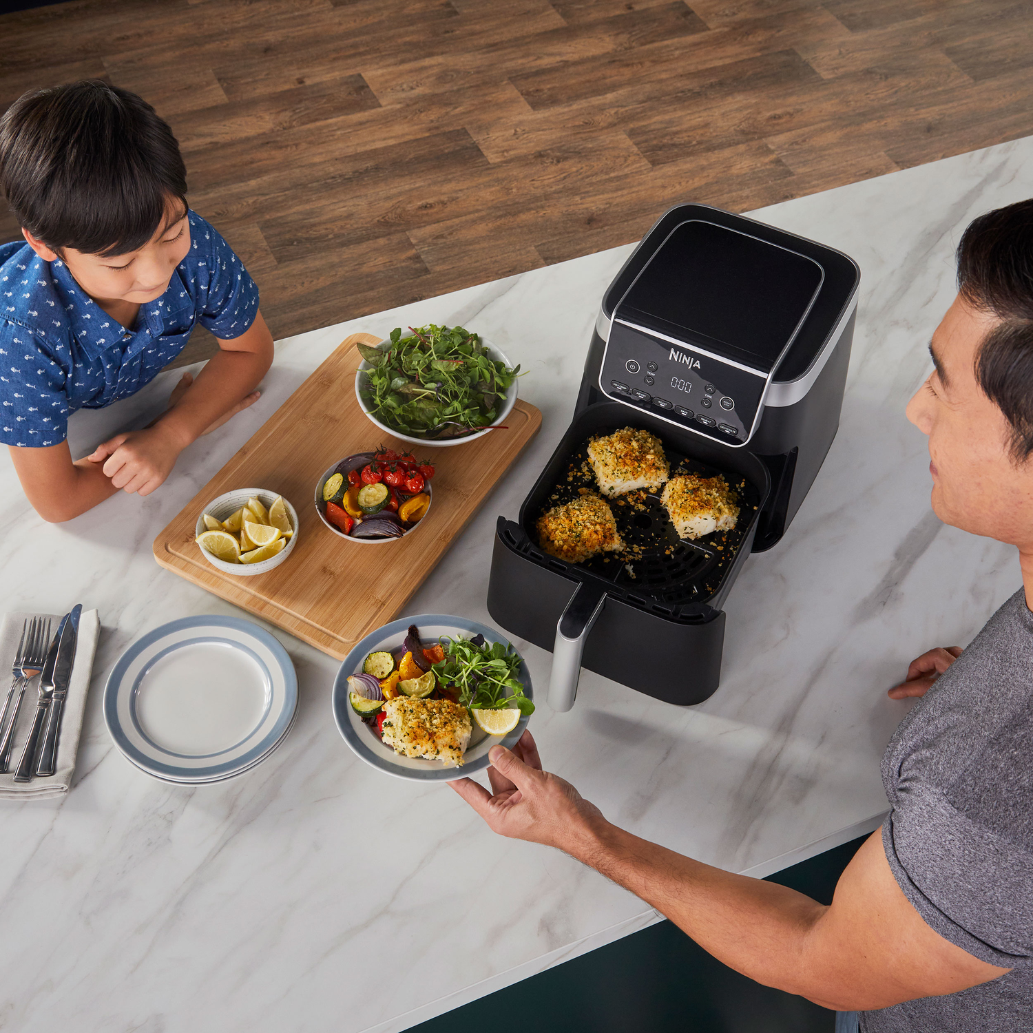 A man serves a plate of food, including fish from a Ninja air fryer. A boy watches. Other plates and a wooden cutting board are present.