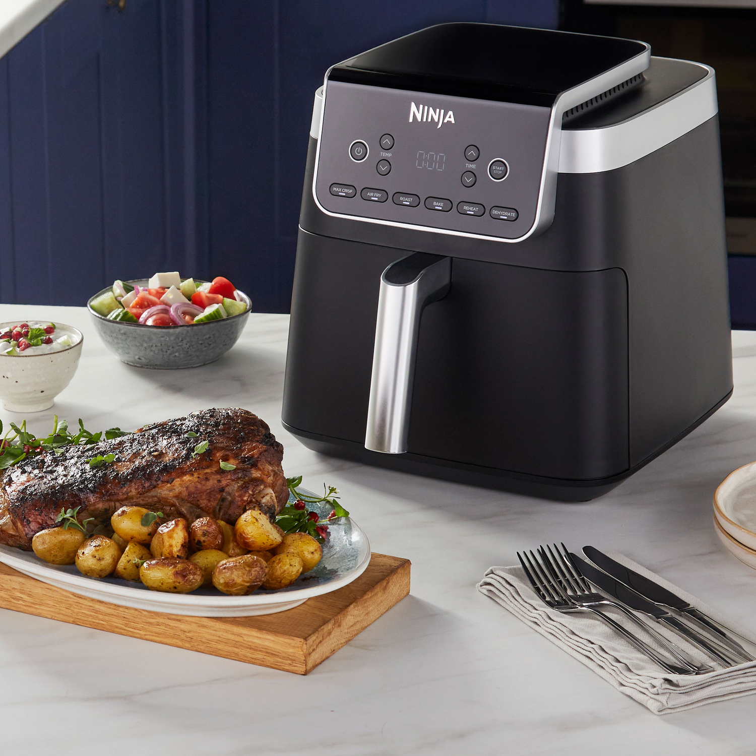 A black Ninja air fryer on a marble countertop. A platter of roasted meat and potatoes, salad, and silverware are present.