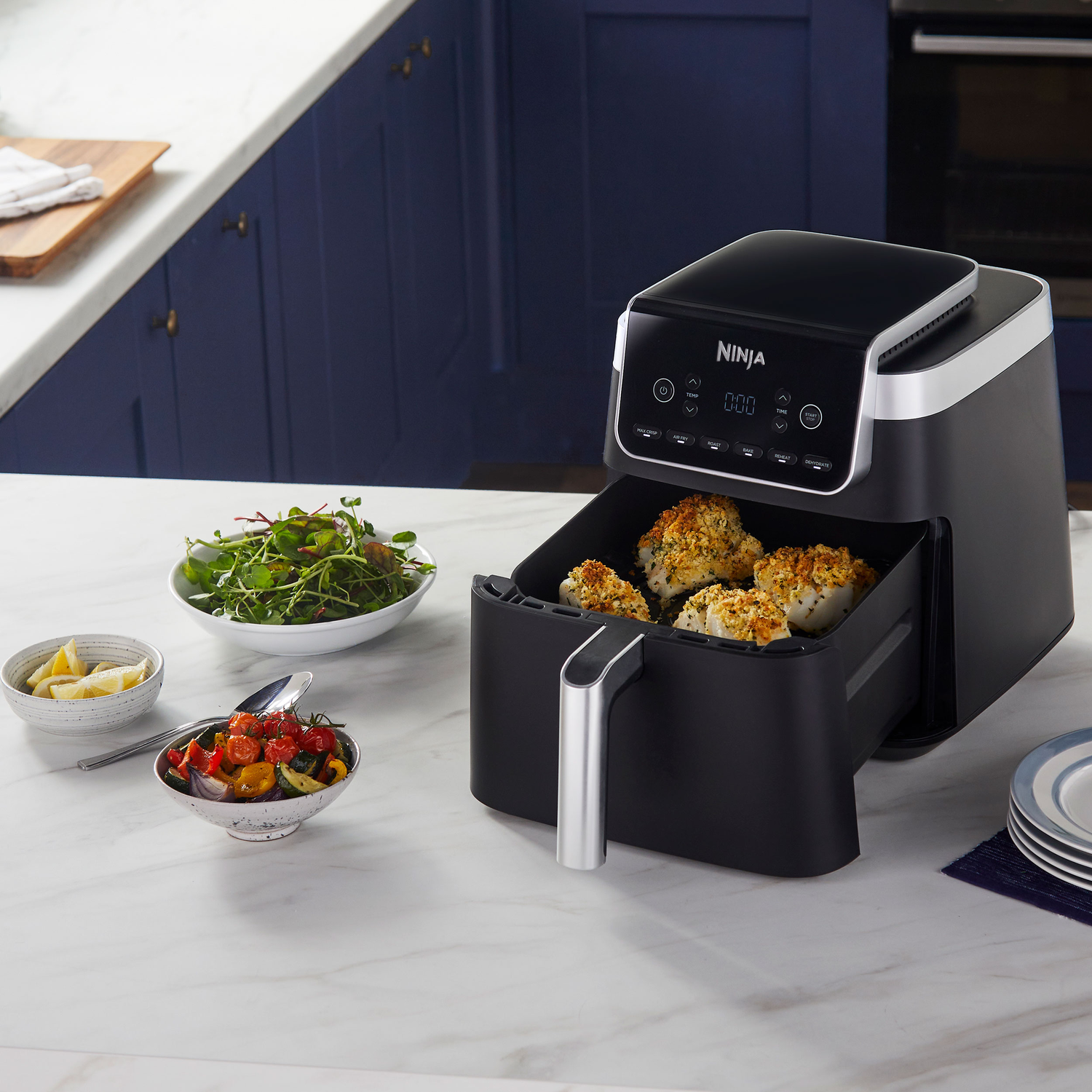 Black Ninja air fryer with cooked food, bowls of salad and vegetables, and a stack of plates on a white countertop.