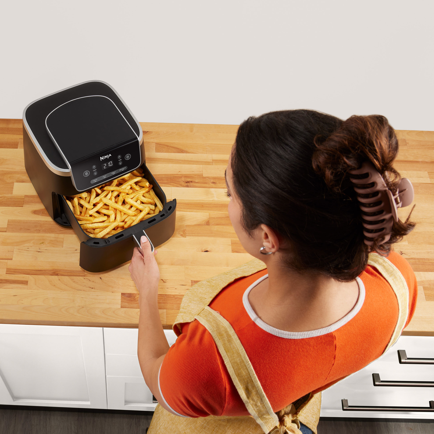 Vrouw die een airfryer gebruikt. Airfryer is zwart. Frietjes erin. Houten aanrecht en een witte kast.