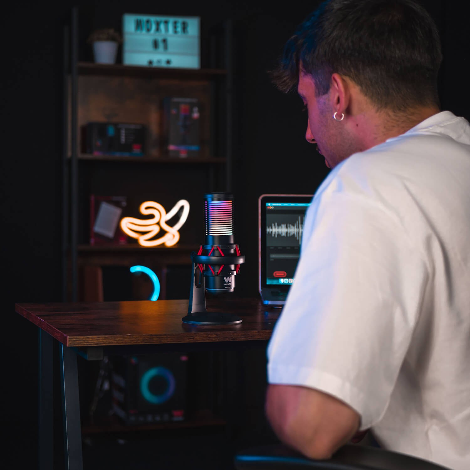 Hombre usando micrófono, tableta en una mesa. Luces de neón y estanterías detrás.