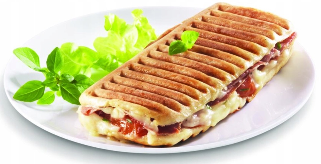 Grillowana kanapka Panini na białym talerzu z sałatą. Chleb ma ślady po grillu, wypełniony serem, mięsem i bazylią.