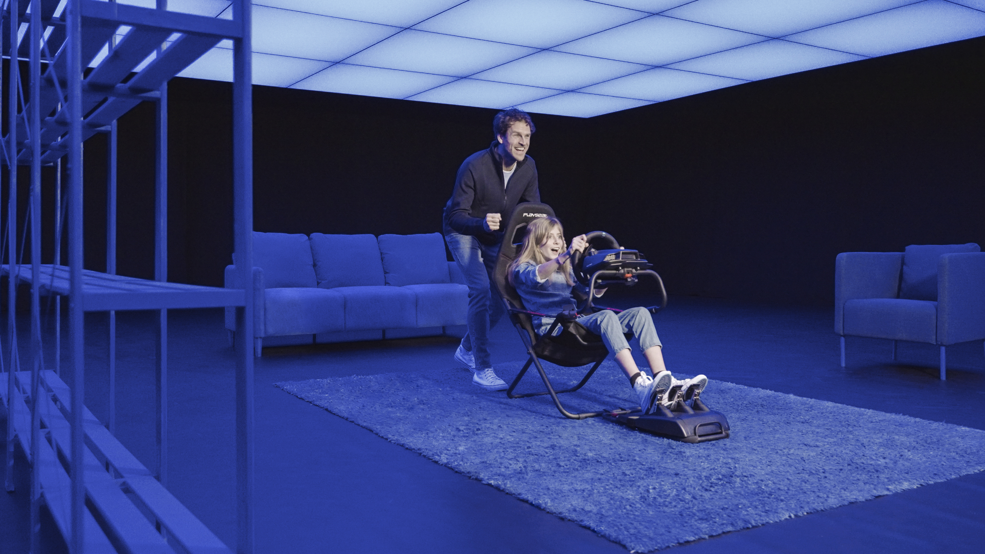 Ein Mann schiebt ein Mädchen in einem Rennsimulatorsessel. Blau beleuchteter Raum mit Sofa und Regalen.