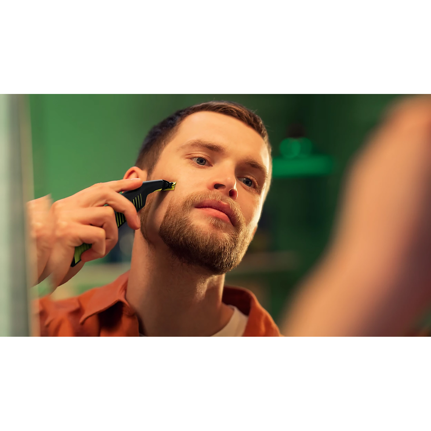Un uomo si rade il viso con un regolabarba Philips nero e verde in uno specchio.