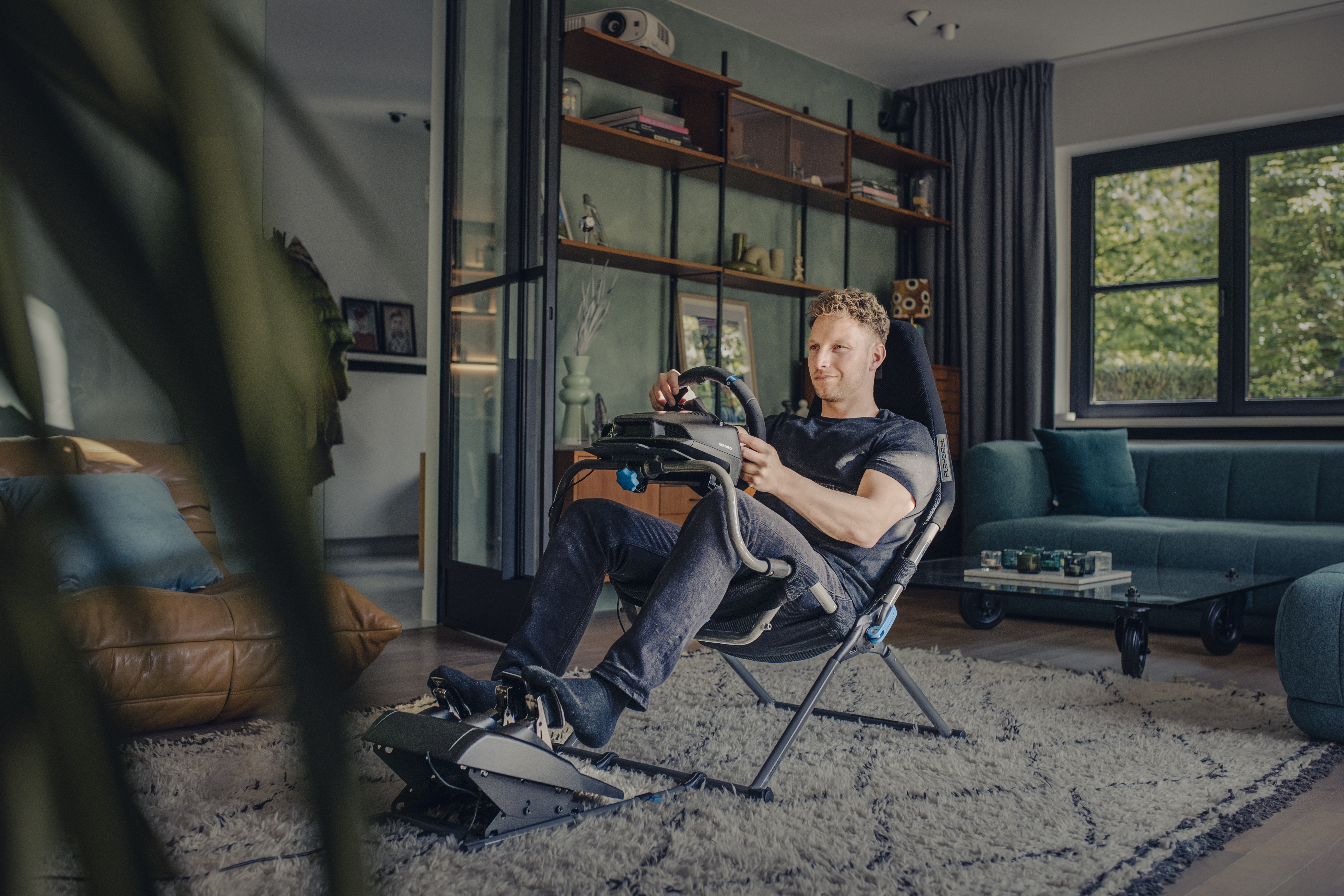 Mann in einem Wohnzimmer, der mit Lenkrad und Pedalen ein Rennspiel spielt.