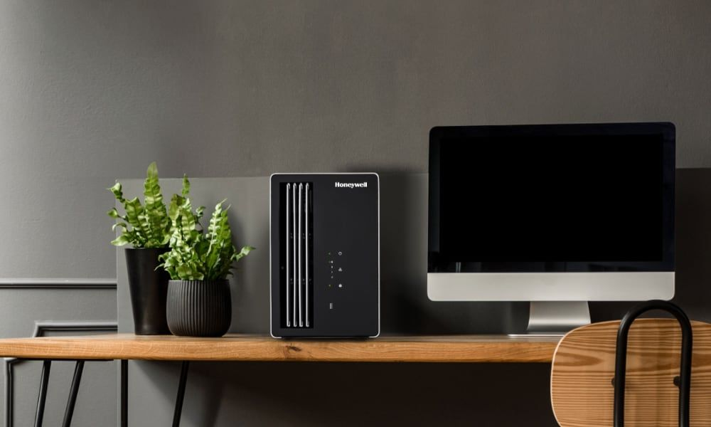 Czarny oczyszczacz powietrza na drewnianym biurku. Monitor komputera, rośliny i drewniane krzesło. Szara ściana.