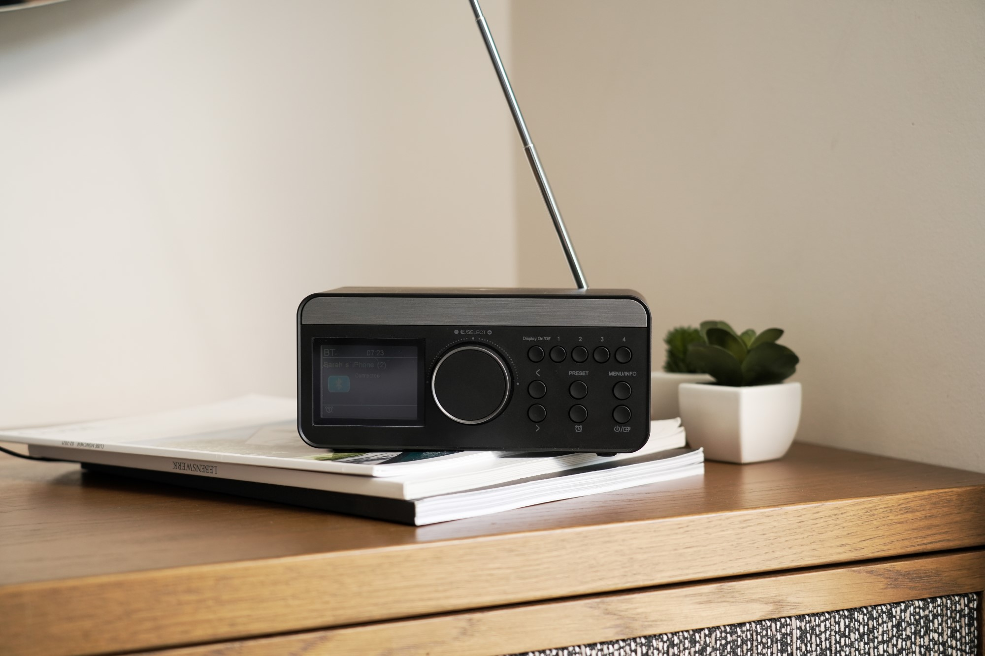 Czarny radioodbiornik na drewnianej powierzchni, z małą rośliną i czasopismami. Radio ma wyświetlacz.