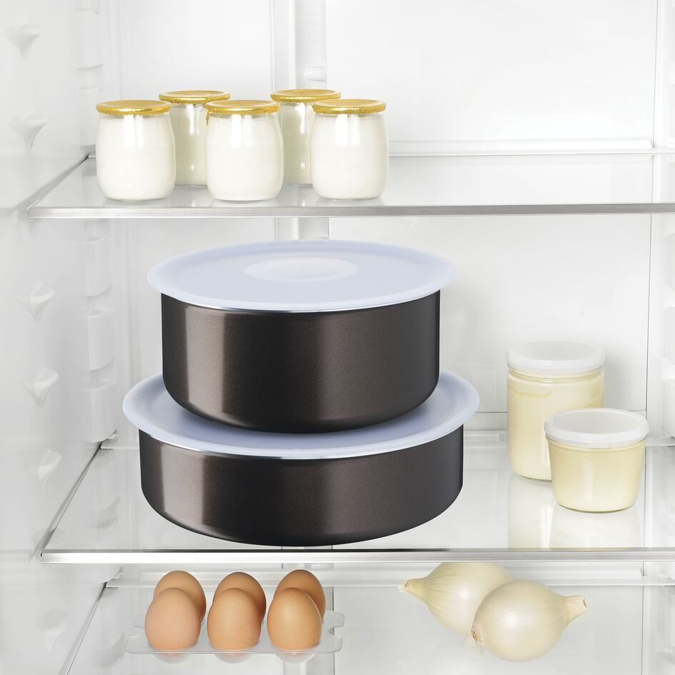 Inside a refrigerator, two black bowls with white lids, jars, and eggs, with onions on the bottom.