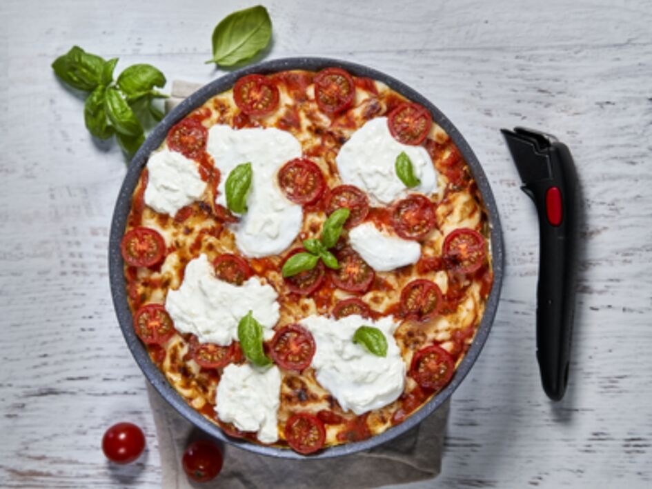Pizza in a pan with tomatoes, mozzarella, and basil, next to a black pizza cutter.