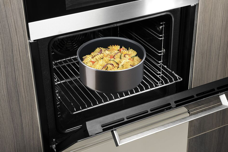 An oven with the door open, showing a round pan with pasta and vegetables inside.