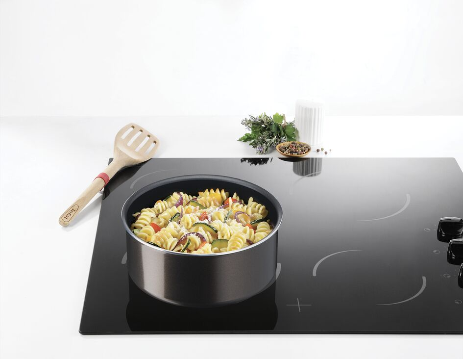 Pasta in a pot on a black induction cooktop. Other ingredients and a wooden spoon are nearby.