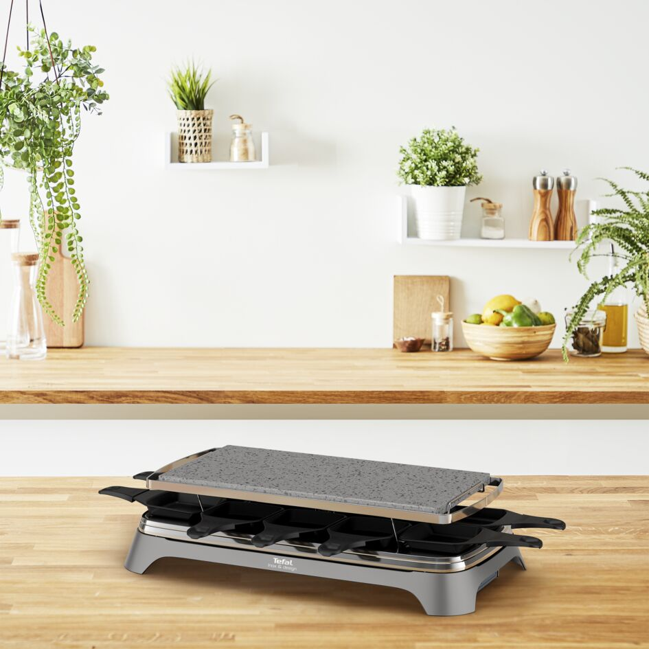 Een raclette grill op een houten tafel. De achtergrond heeft planken en planten.