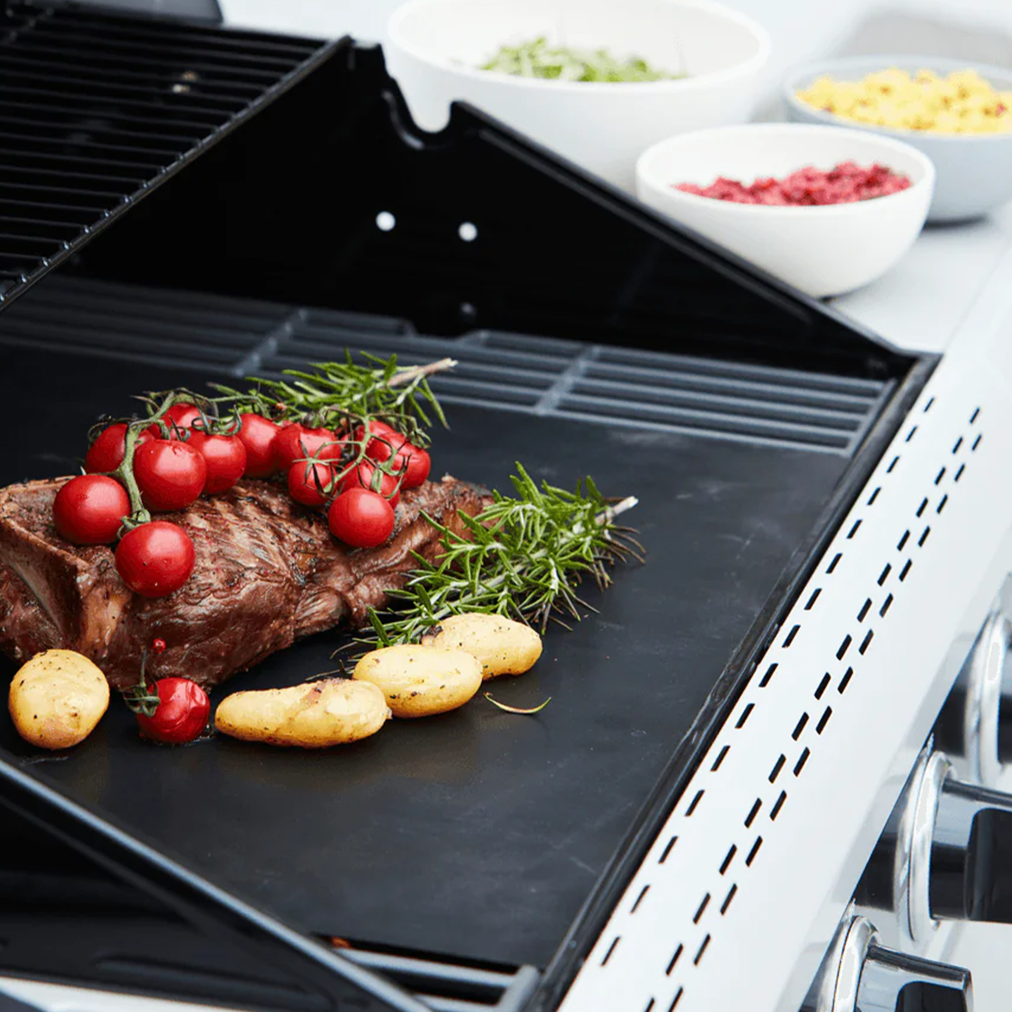 Egy steak paradicsommal és burgonyával a grillen. A háttérben tálak vannak.
