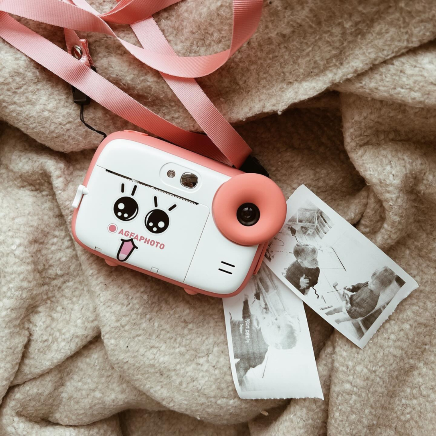Pink and white toy camera with photos on a soft, beige blanket.