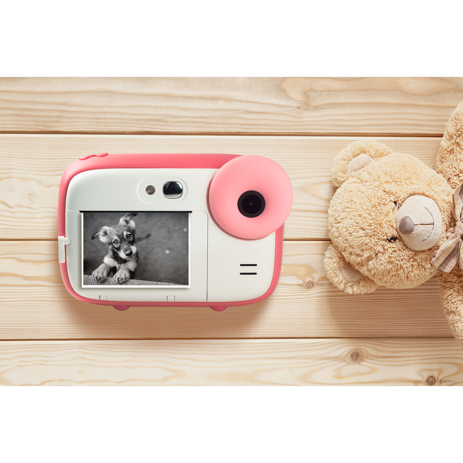 A pink and white instant camera displays a puppy photo. A teddy bear sits on a light wood surface.