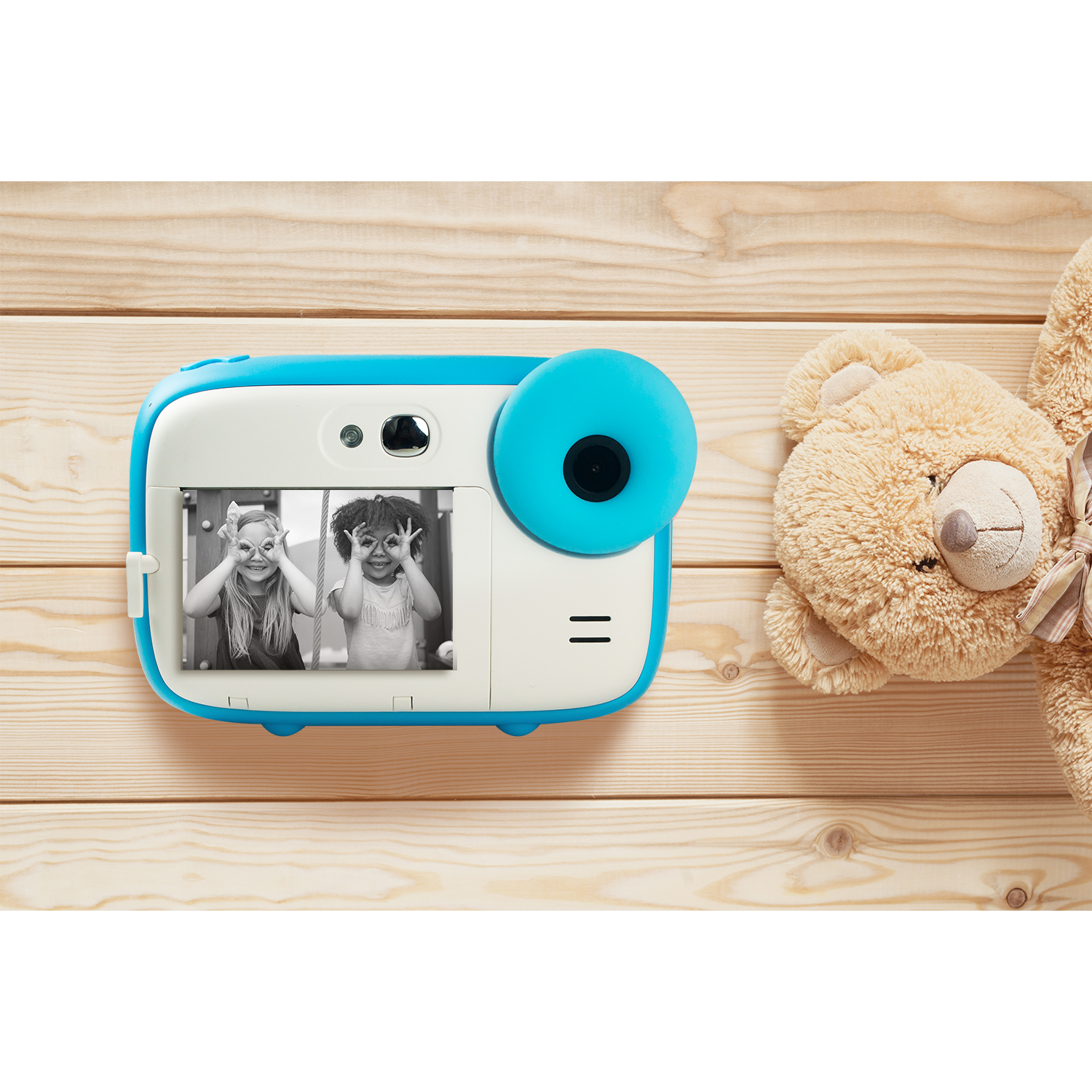 Blue and white instant camera with a photo of two girls making faces, next to a teddy bear.