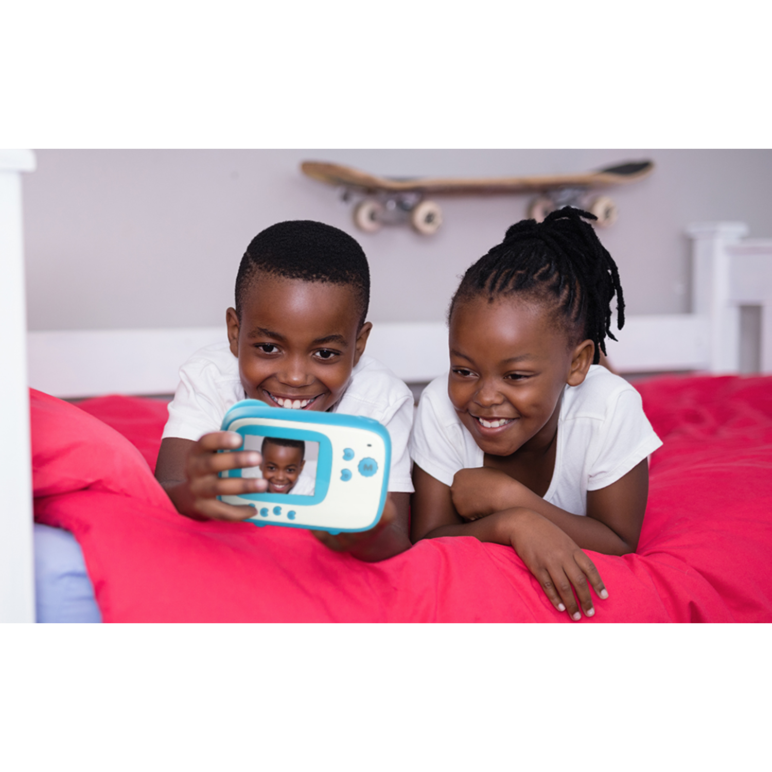 Two smiling children taking a selfie with a blue and white camera, lying on a red bed.