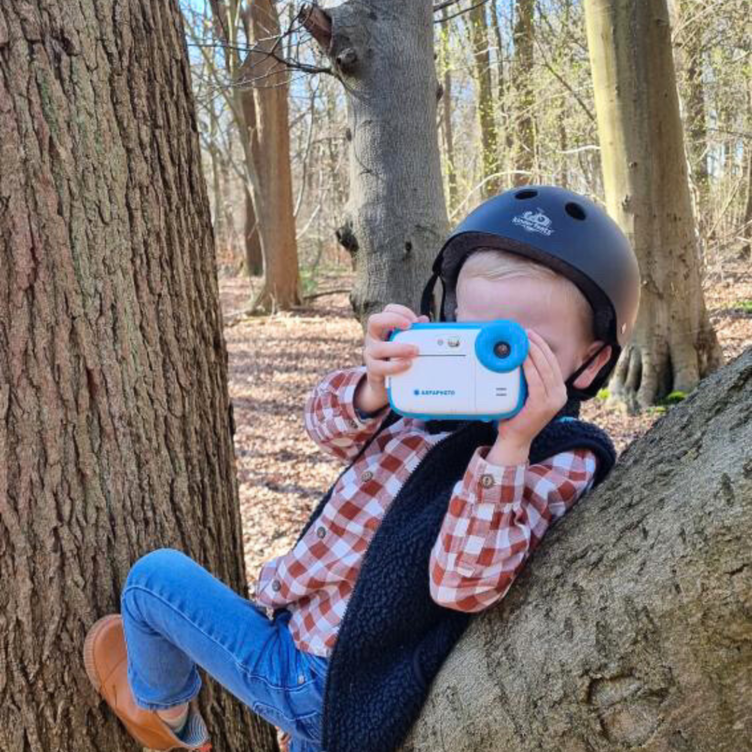 A child in a plaid shirt and helmet taking a picture with a blue camera in a forest.
