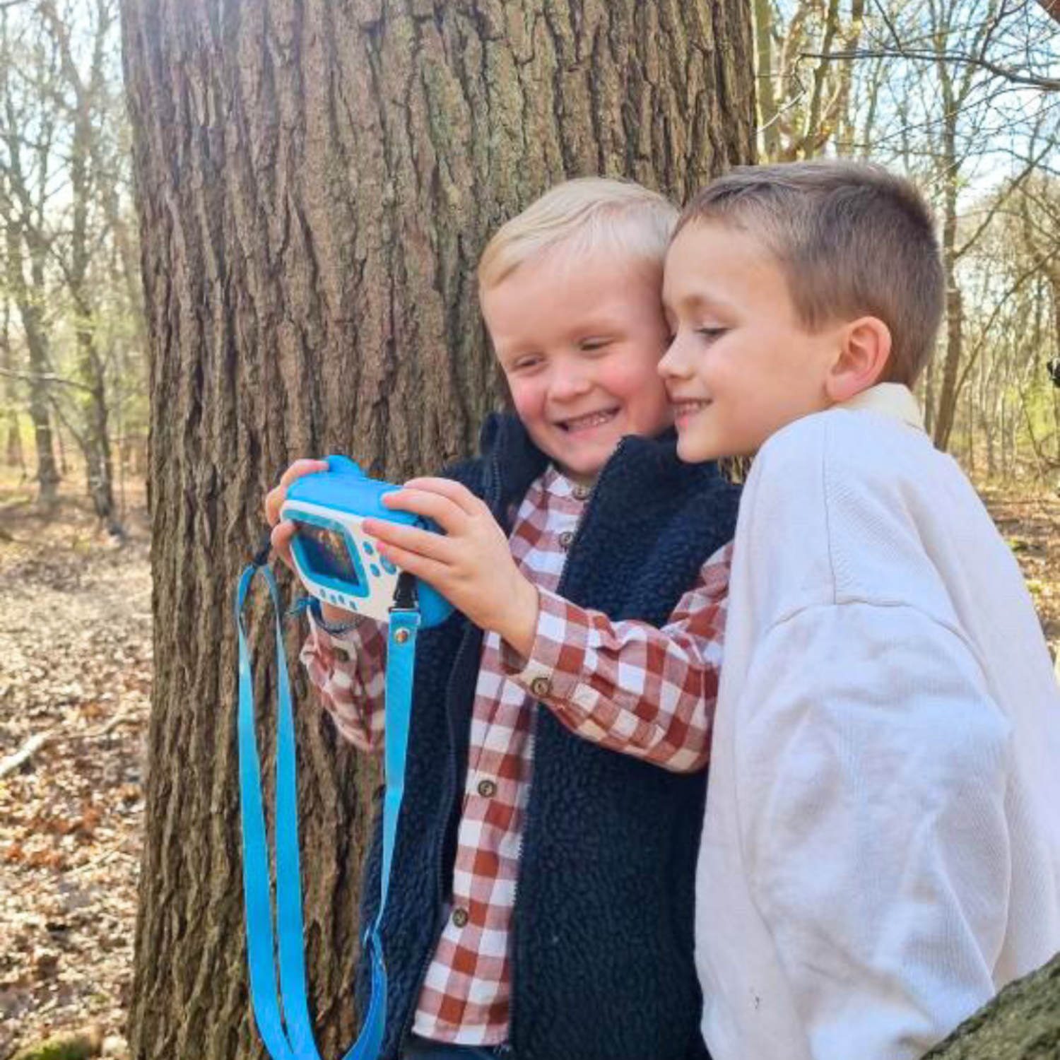 Two smiling boys looking at a blue and white camera near a tree trunk in the forest.