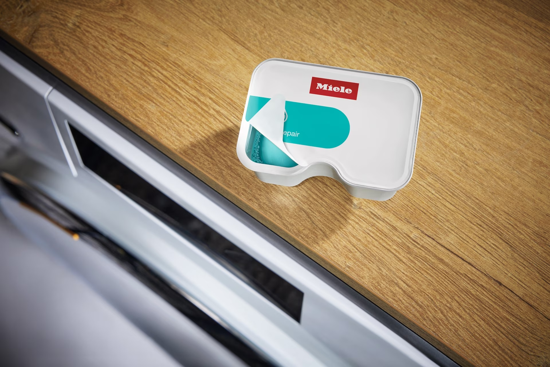 Biały pojemnik z turkusową tabletką czyszczącą w środku, na drewnie. Marka Miele.