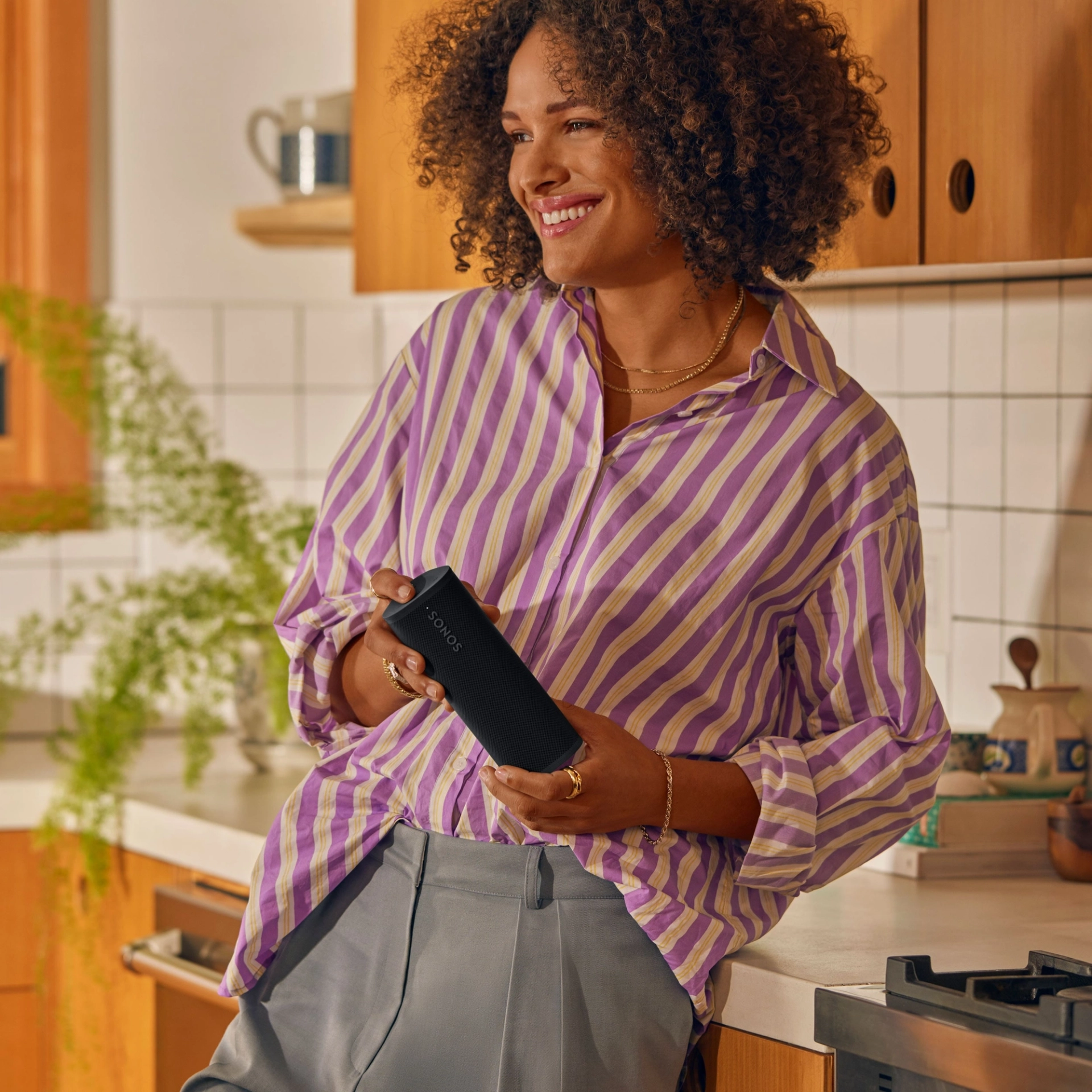 Une femme souriante tenant un haut-parleur noir dans une cuisine, portant une chemise rayée et des bijoux en or.