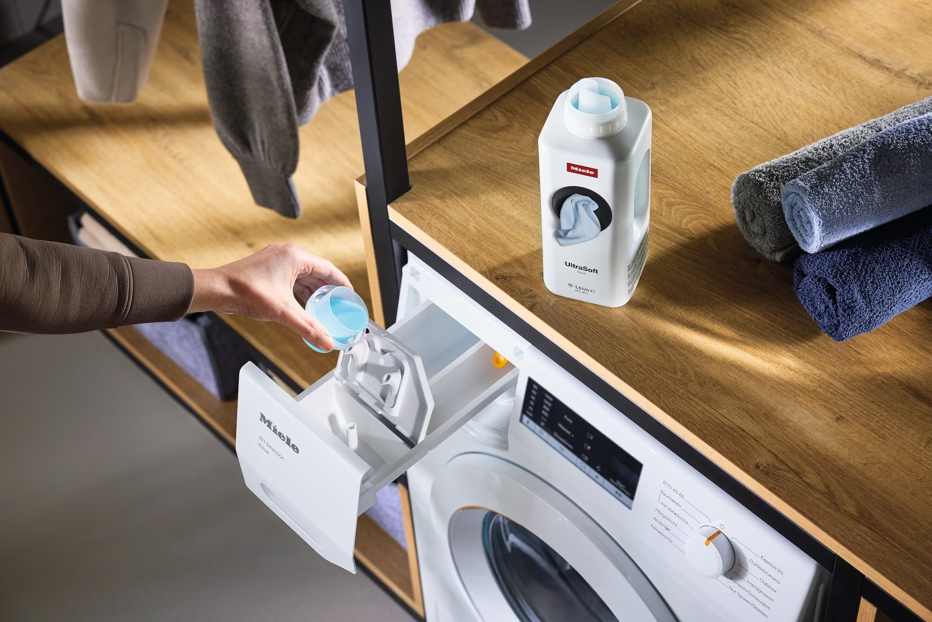 Ręka wlewa detergent do pralki, obok znajdują się złożone ręczniki.