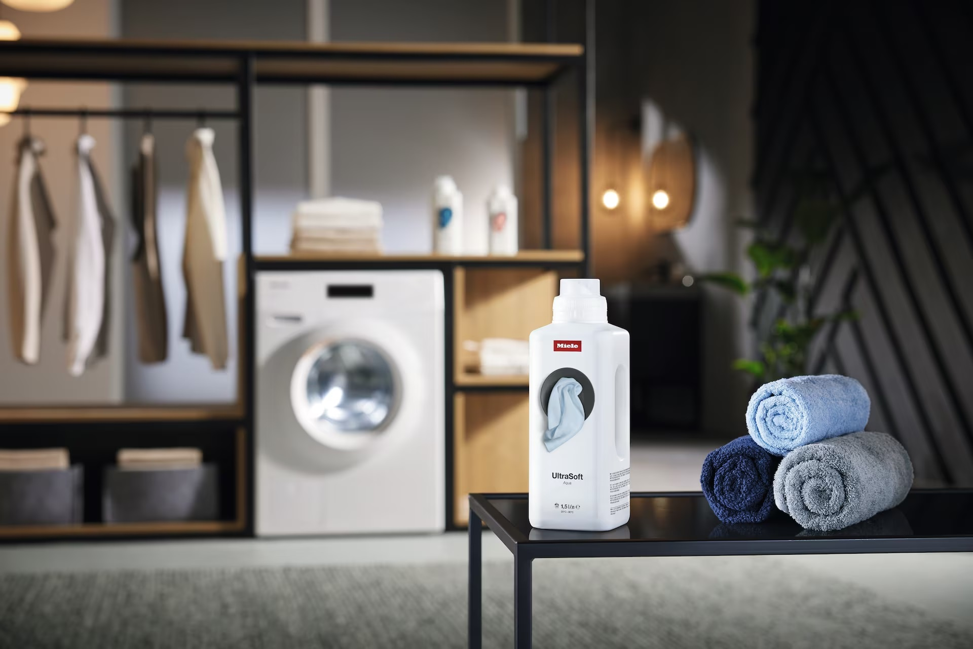 Biała pralka, butelka Miele UltraSoft Aqua i trzy zwinięte ręczniki.