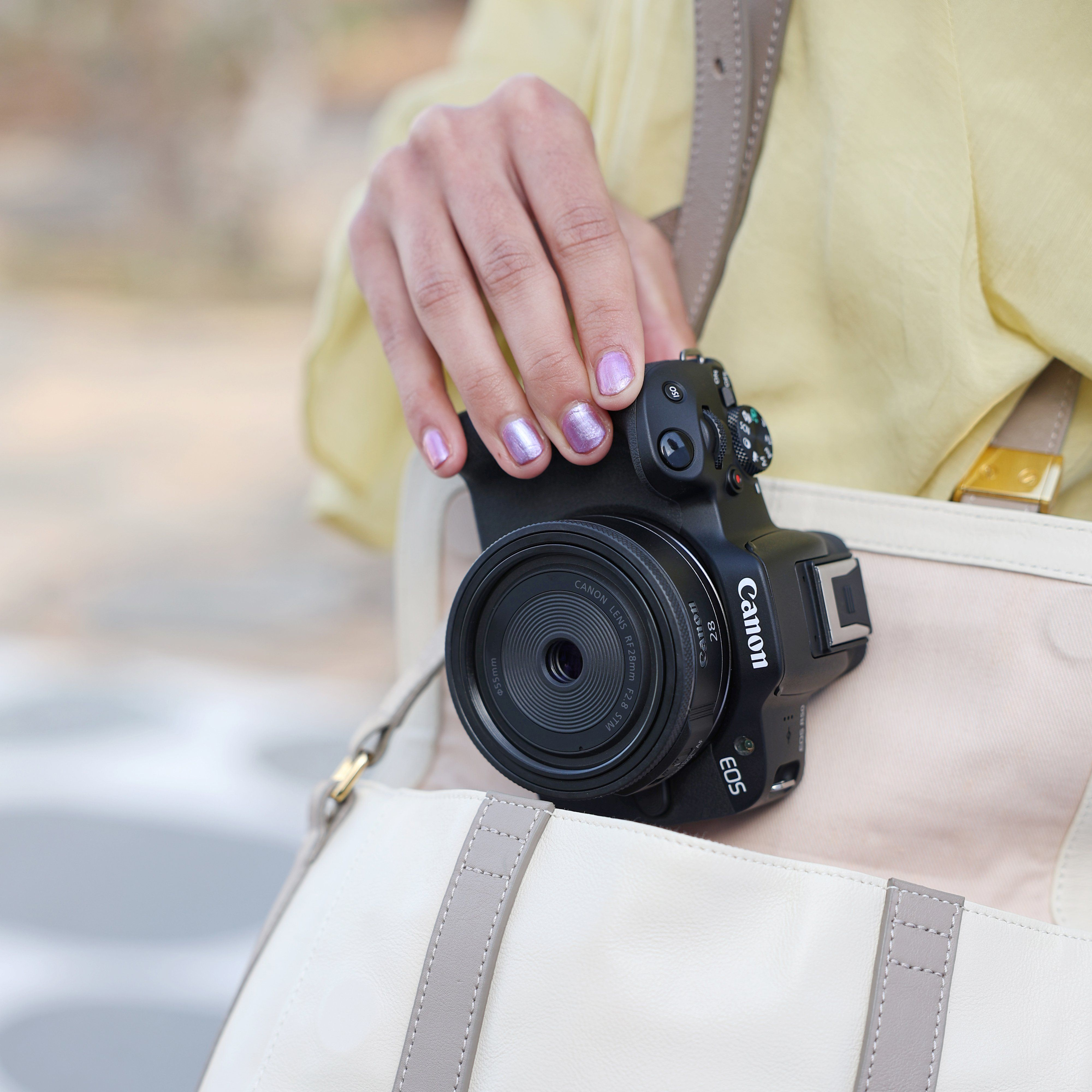 Une main avec du vernis violet tient un appareil photo Canon noir sur un sac crème.