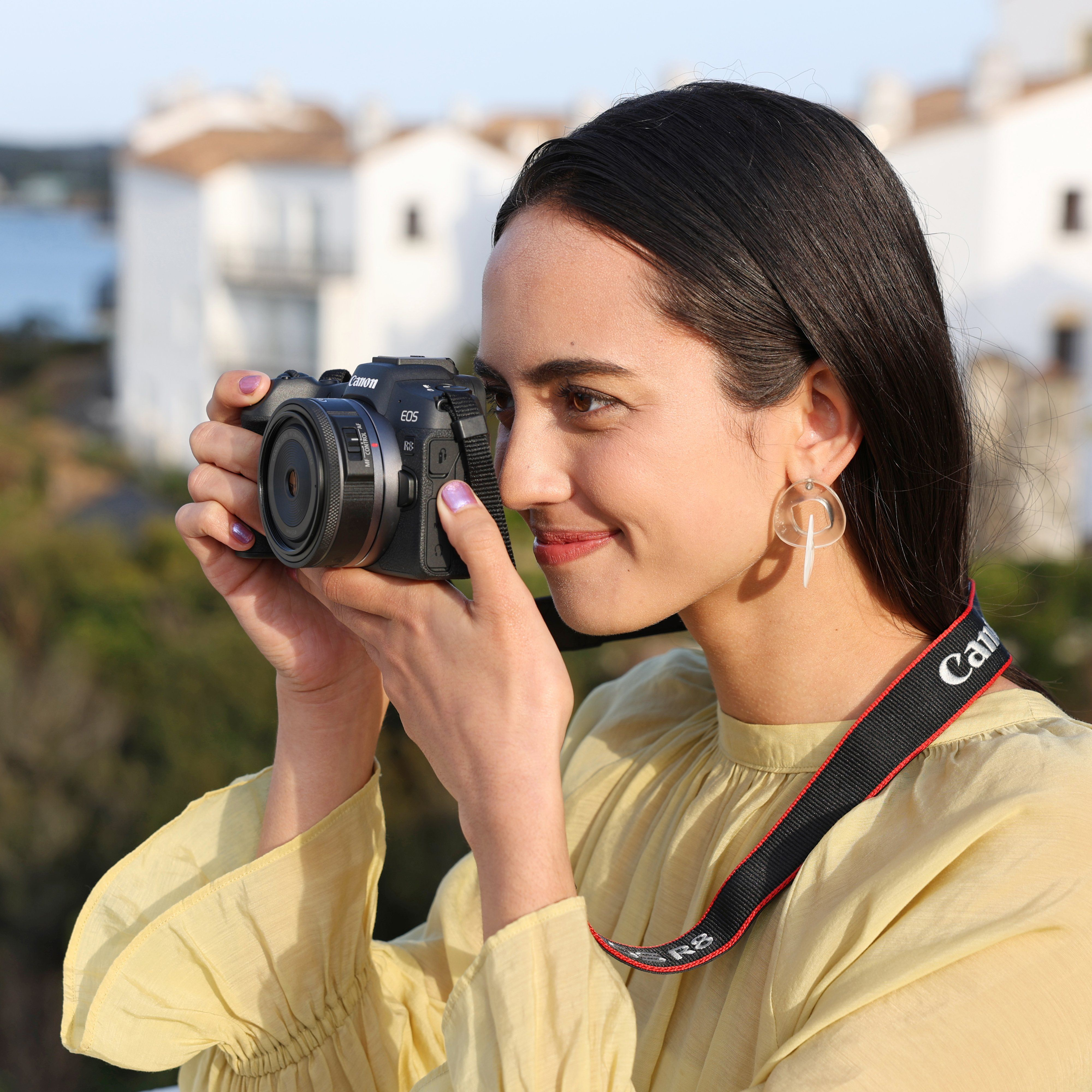 Une femme aux cheveux sombres tient un appareil photo, regardant vers la droite. Elle porte un haut jaune.