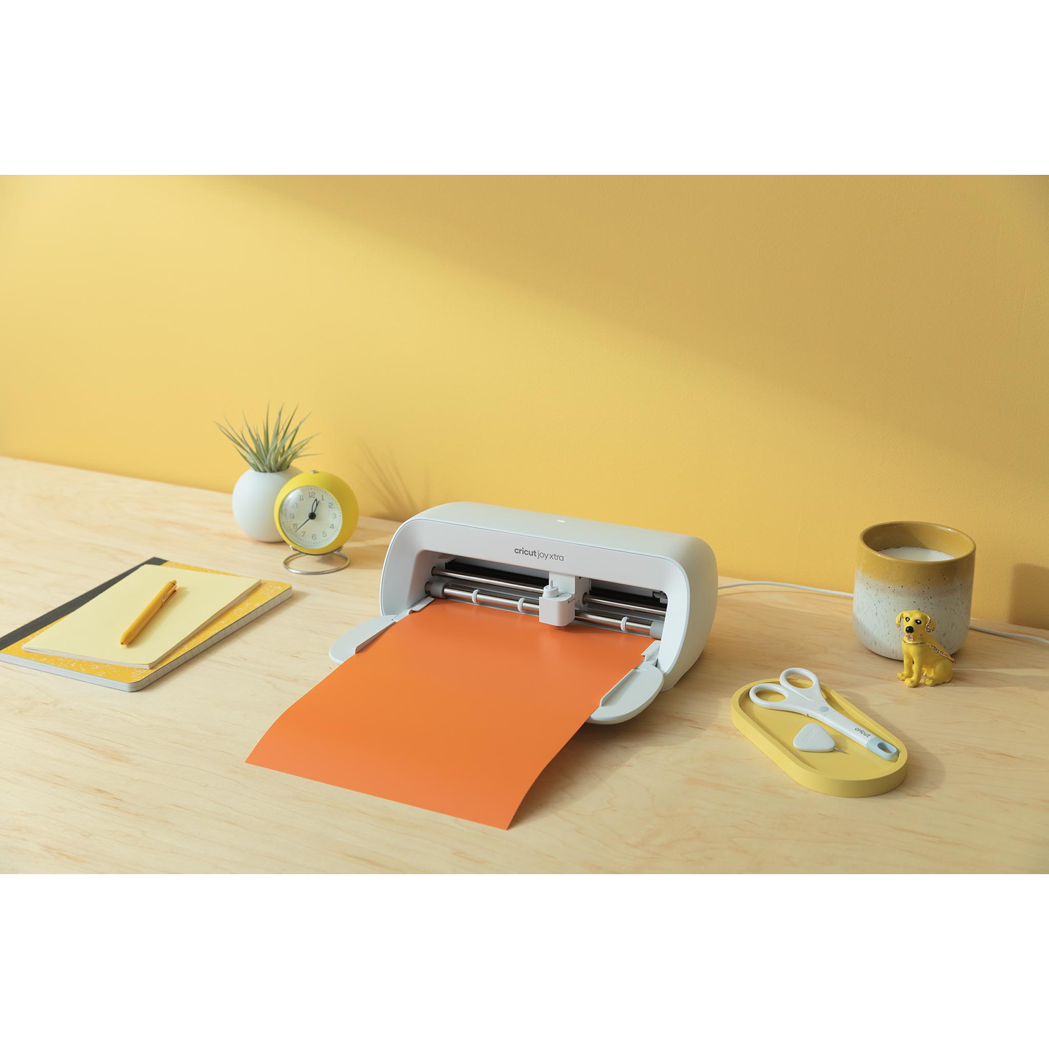 Een witte snijmachine met oranje papier dat erin wordt gevoerd. Er is een gele klok en andere items.