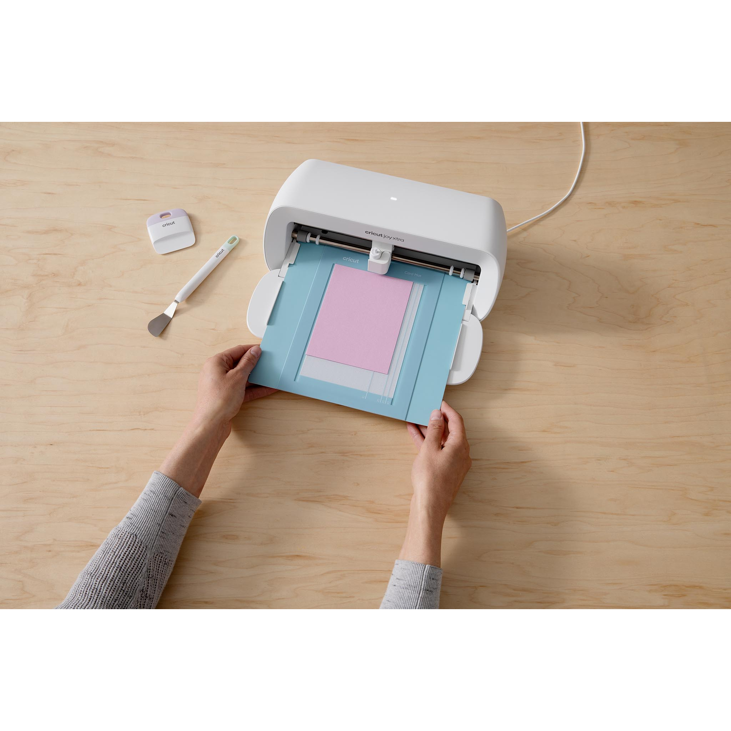 Hands placing pink paper into a Cricut cutting machine on a wooden surface. Tools are nearby.