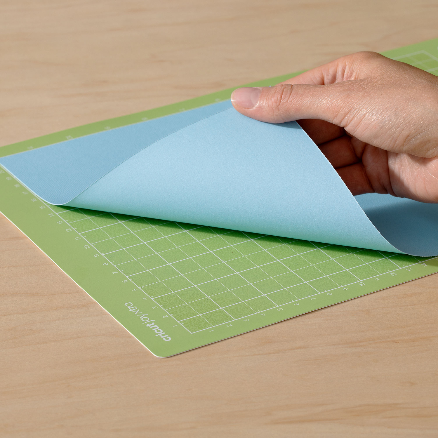 Hand die een blauw vel van een groene snijmat afhaalt. De mat heeft een rasterpatroon en tekst 'Cricut by Xtra'.