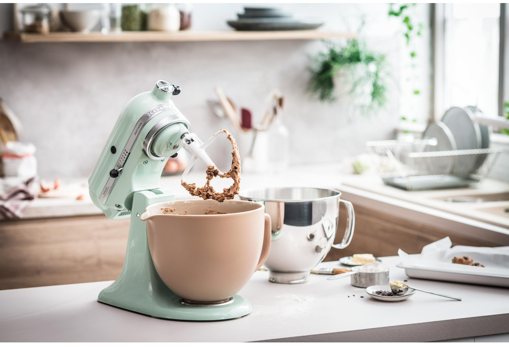 Zöldeskék KitchenAid mixer bézs tálban keveri a tésztát, konyhai környezetben.