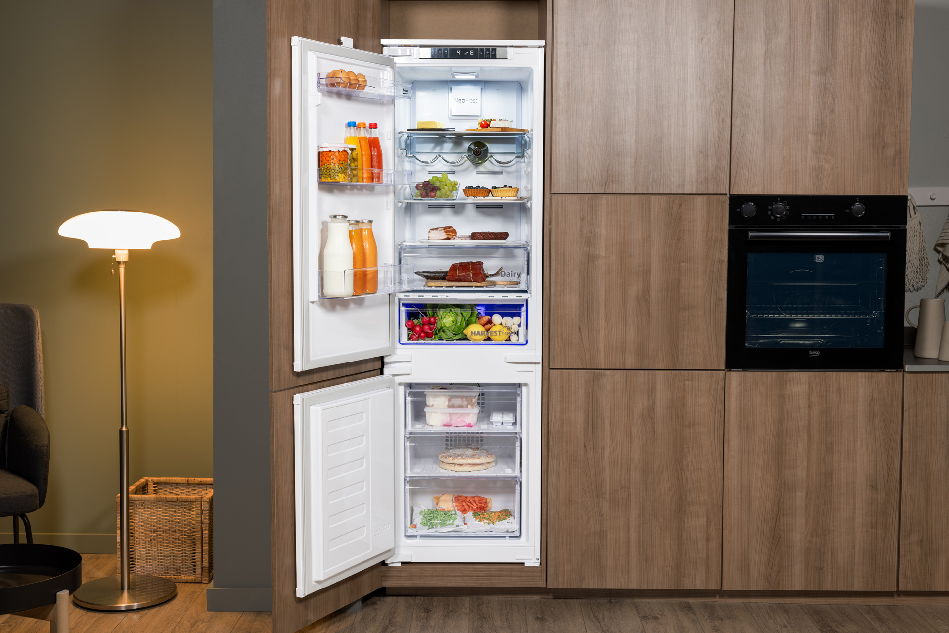 Otwarta lodówka z jedzeniem i napojami. Drewniane szafki i piekarnik w tle. Lampa podłogowa.