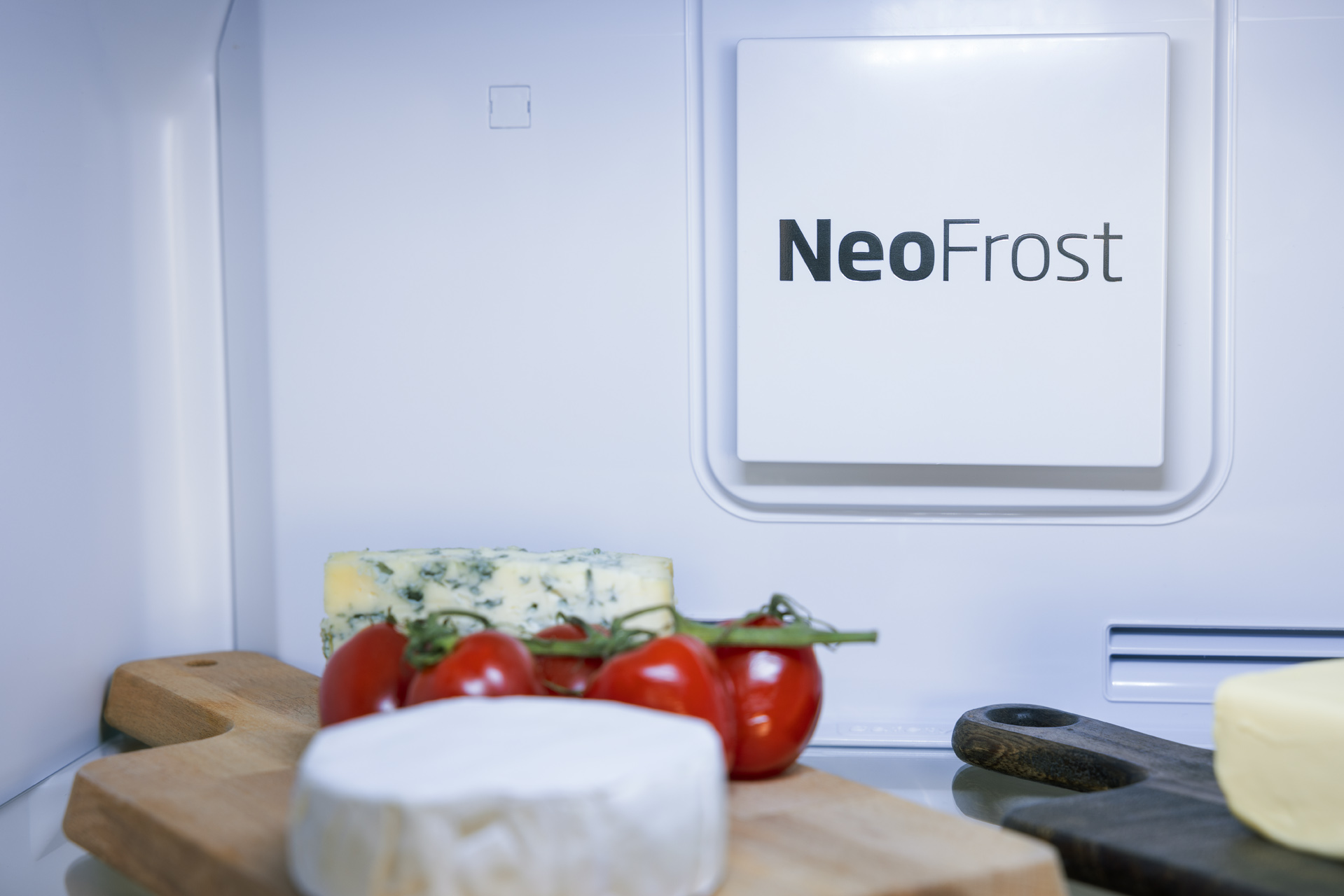 Wnętrze lodówki z etykietą NeoFrost. Widoczne ser, pomidory i drewniana deska do krojenia.
