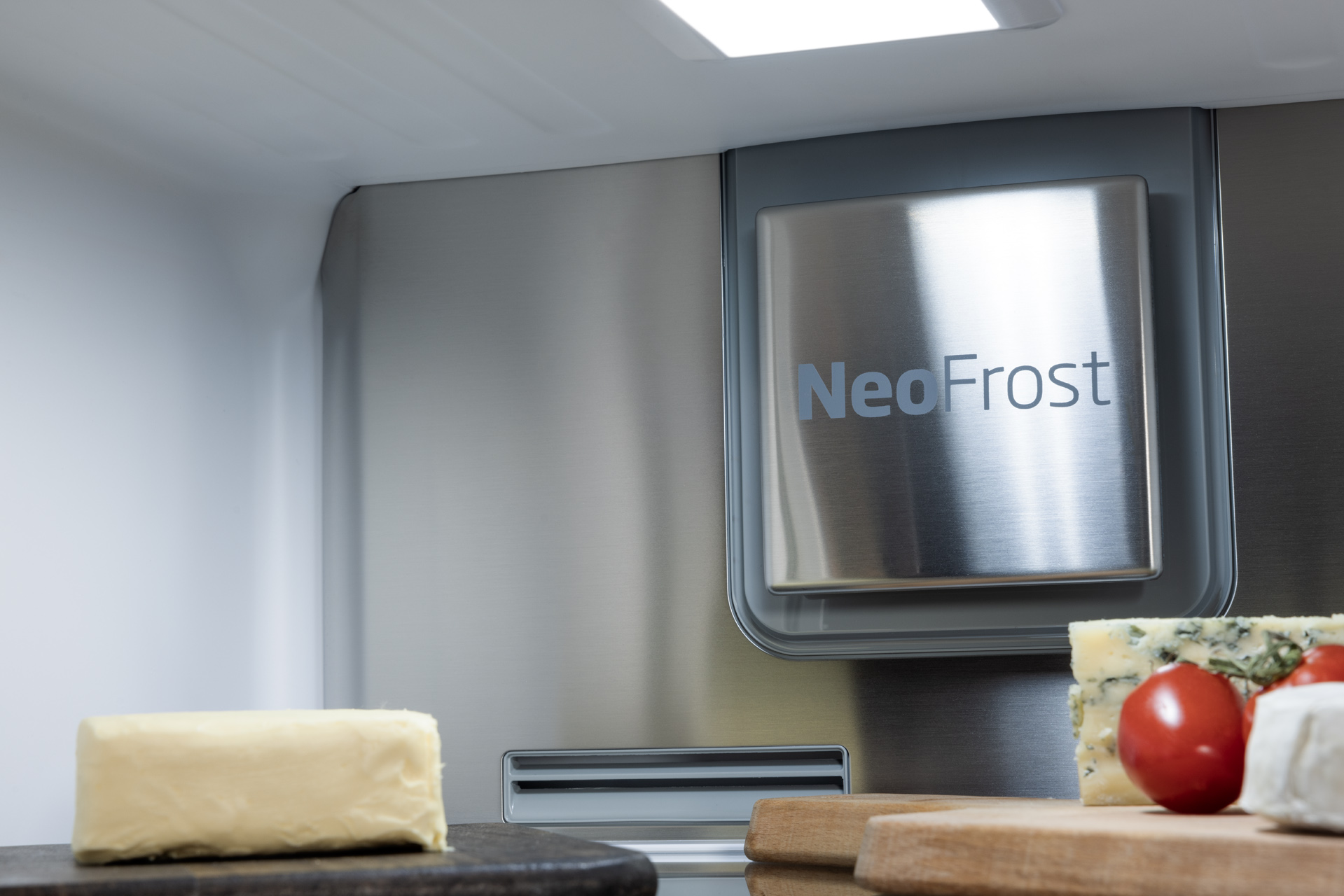 Wewnątrz lodówki z napisem NeoFrost. Ser, pomidor i masło na półce. Srebrne wnętrze.