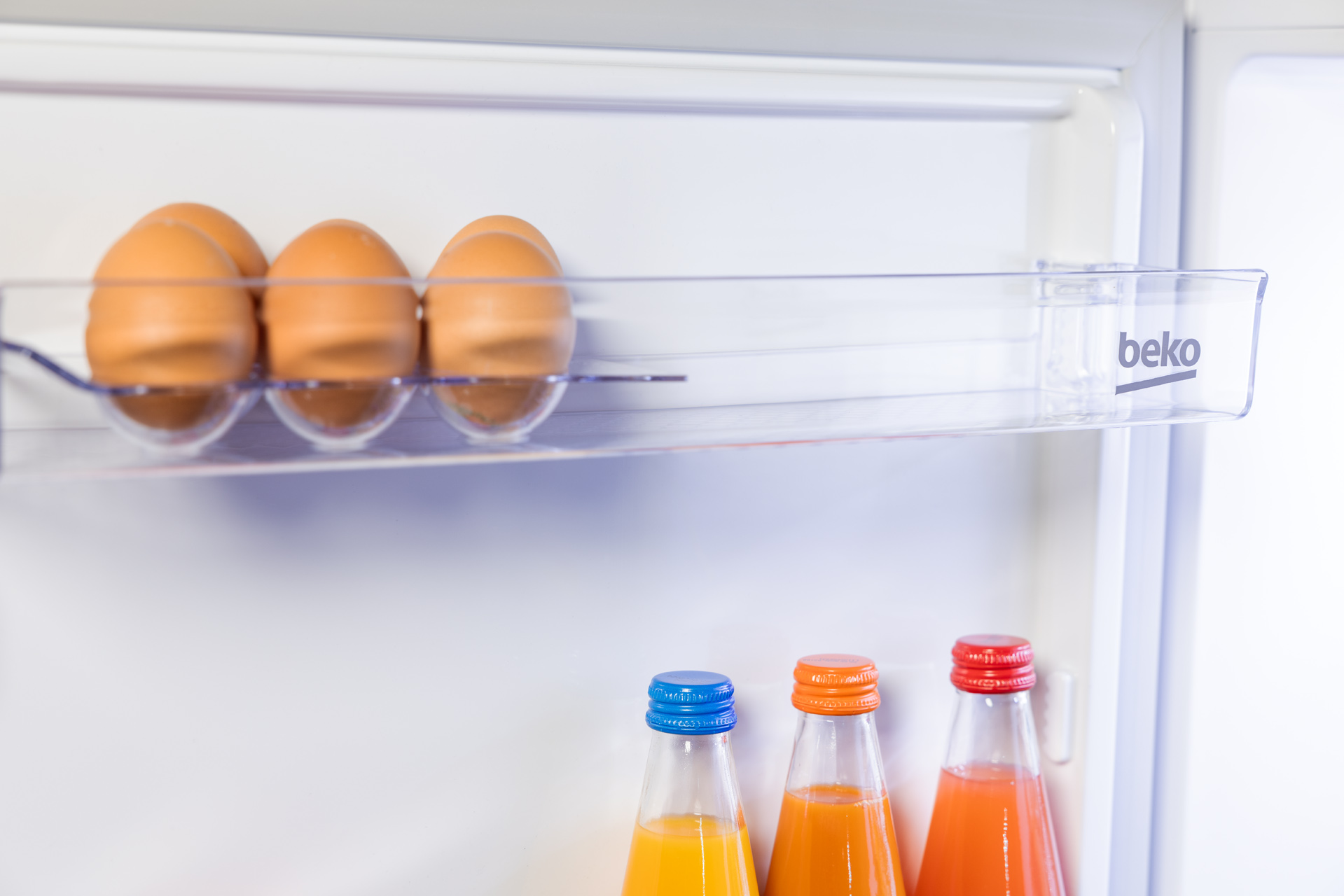 Wewnątrz lodówki, jajka i butelki z sokiem na półkach. Widoczne logo Beko.