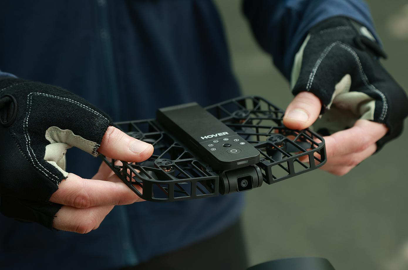 Hände halten eine schwarze Drohne mit Controller, vor verschwommenem Outdoor-Hintergrund.