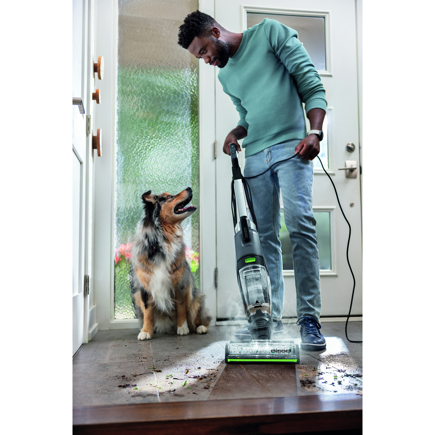 Un uomo passa l'aspirapolvere, guardando un cane. L'aspirapolvere è bianco e nero. Il cane è marrone, nero e bianco.