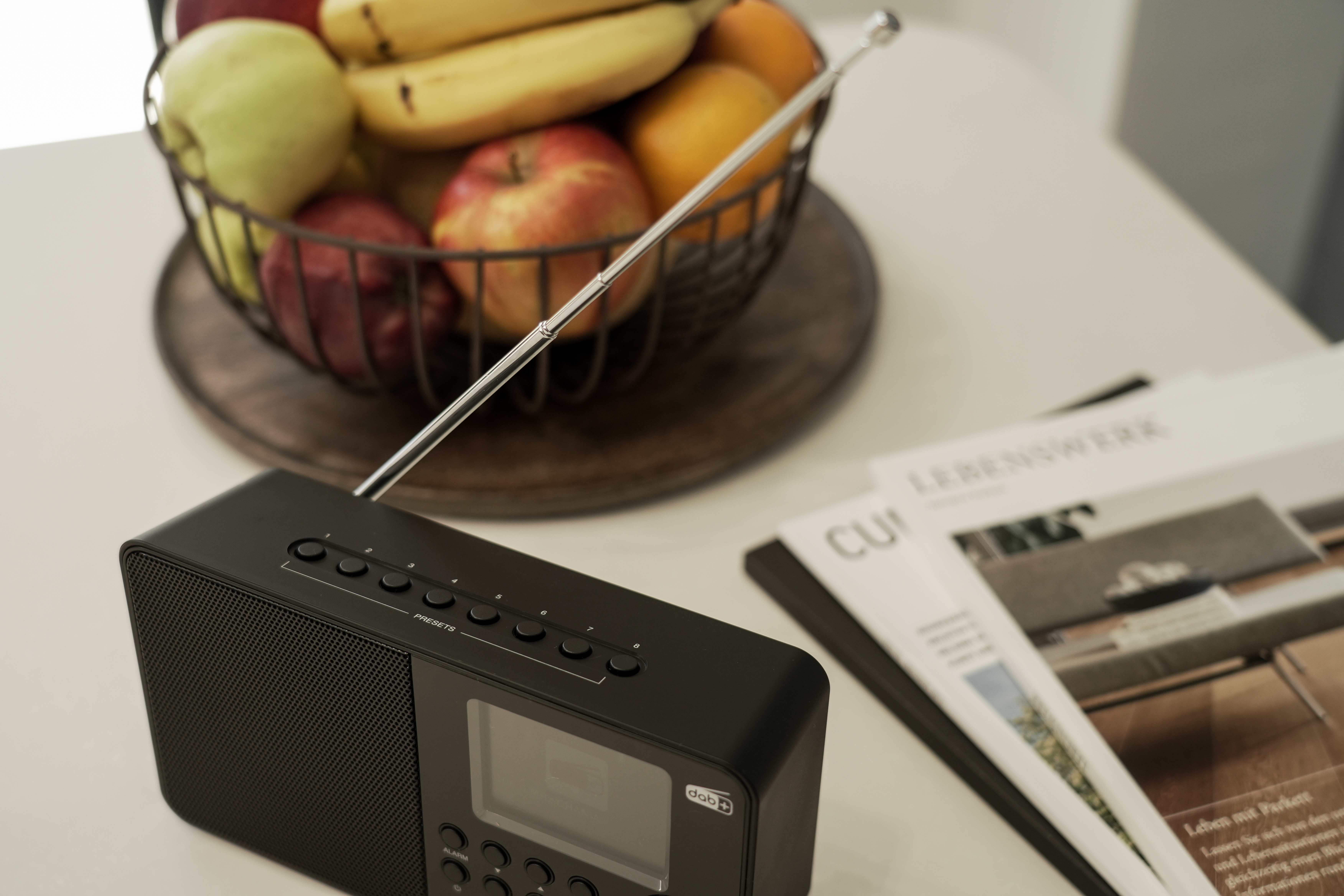 Une radio noire avec des fruits dans un panier et des magazines sur une table blanche.