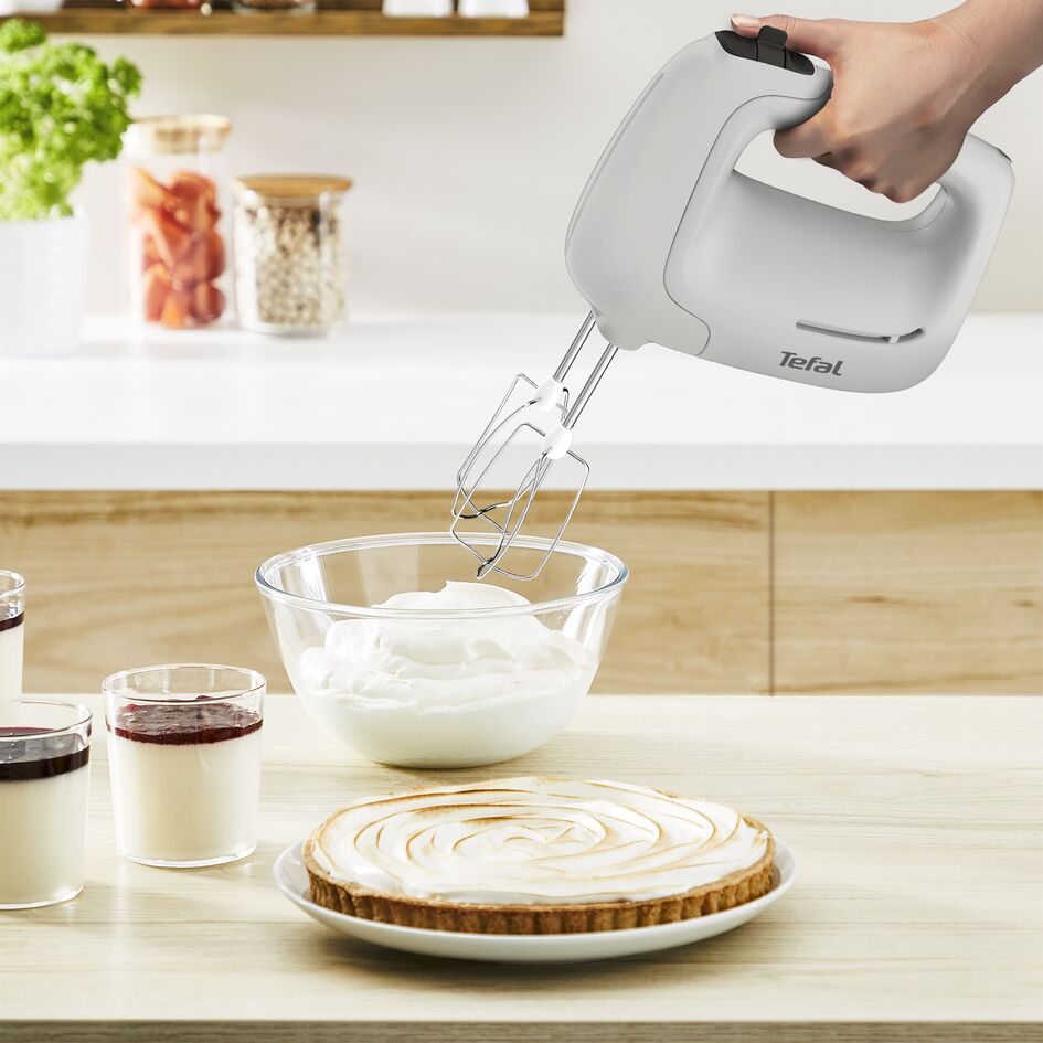 Egy kézi mixer krémet kever egy üvegtálban. Egy torta és desszertpoharak is láthatók.