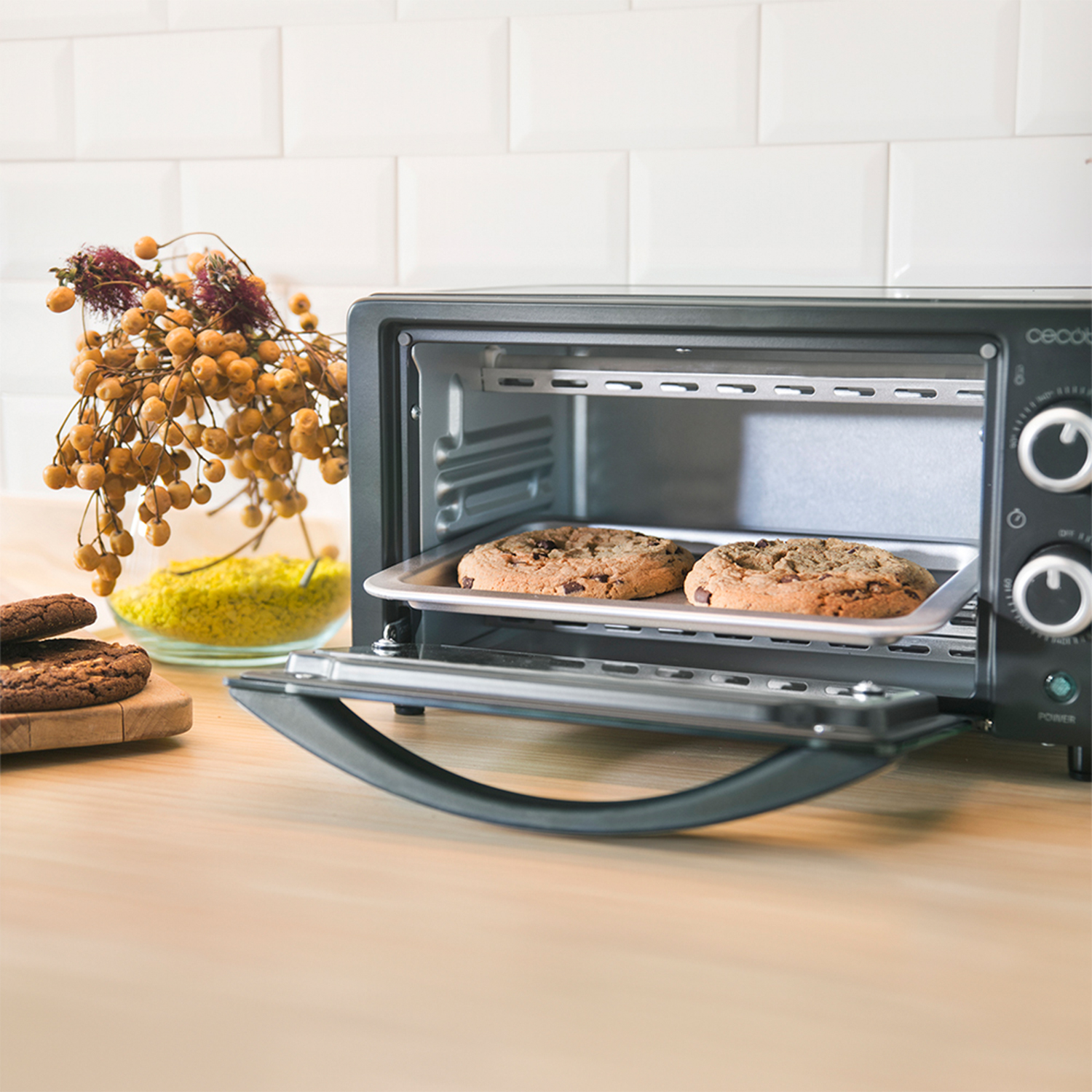 Un forno tostapane nero aperto con due biscotti in cottura. Accanto, ingredienti e decorazioni.