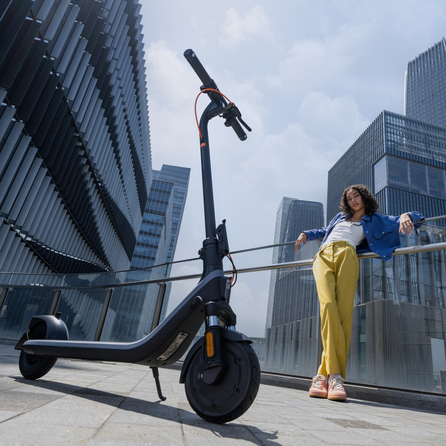 A woman in yellow pants leans against a railing next to a black electric scooter.
