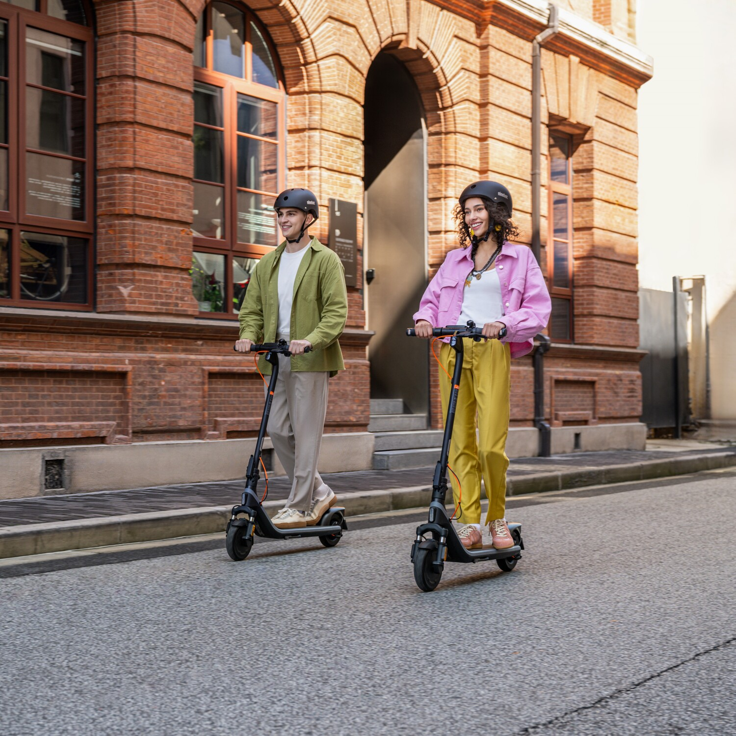 Two people on scooters ride down a street. Brick building in the background.